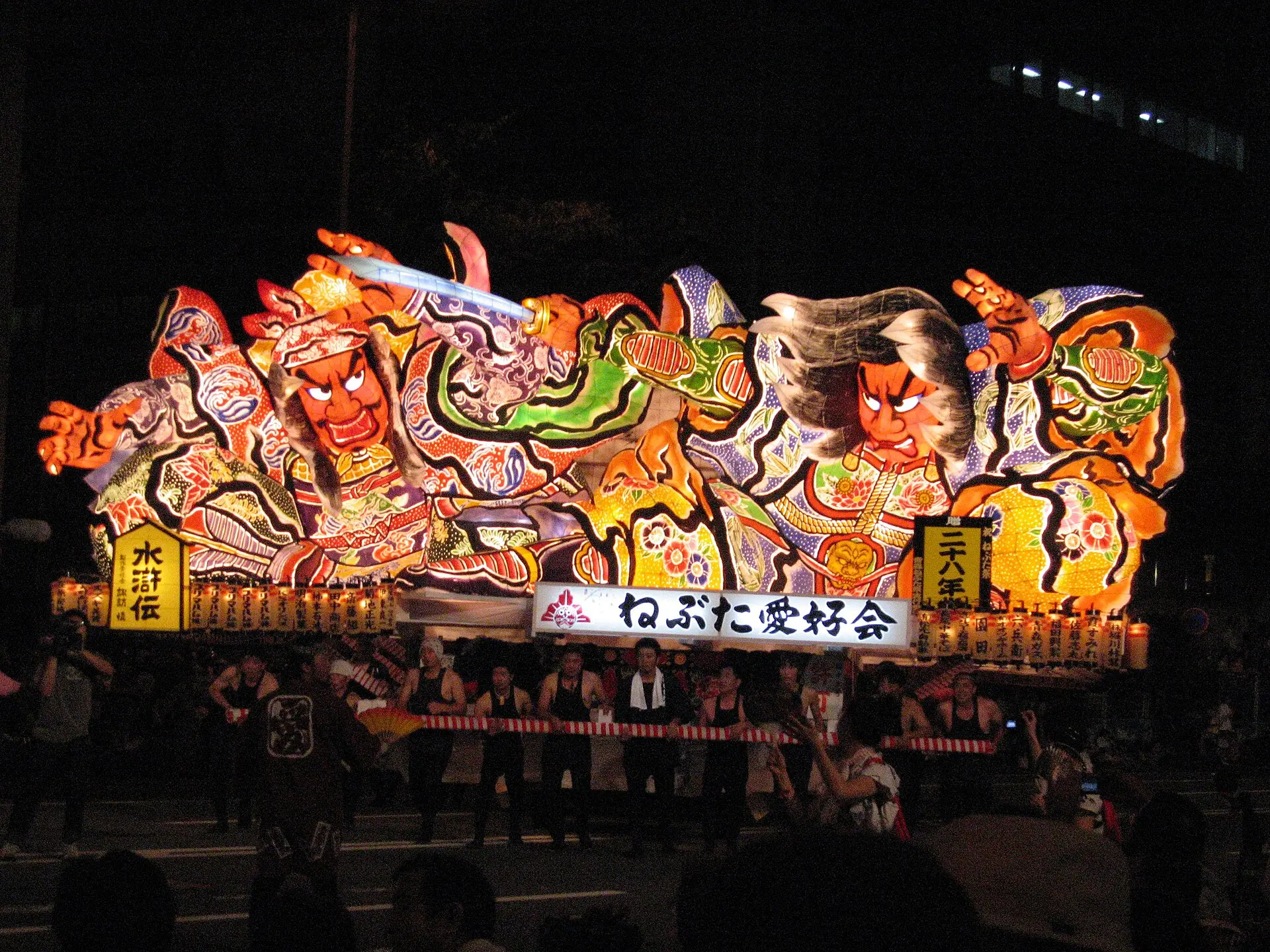夜の大通りを運行する二人の武者を描いたねぶたと曳き手たち
